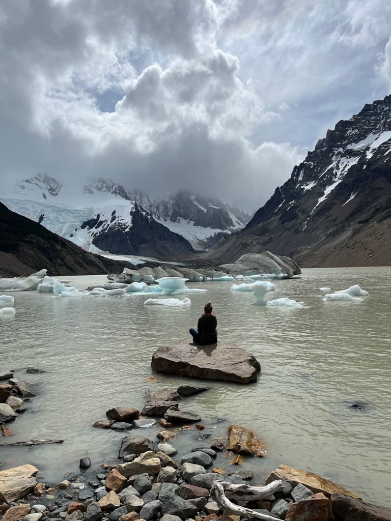 A person sitting on a rock beside an iceberg-filled glacial lake beneath cloudy mountains
