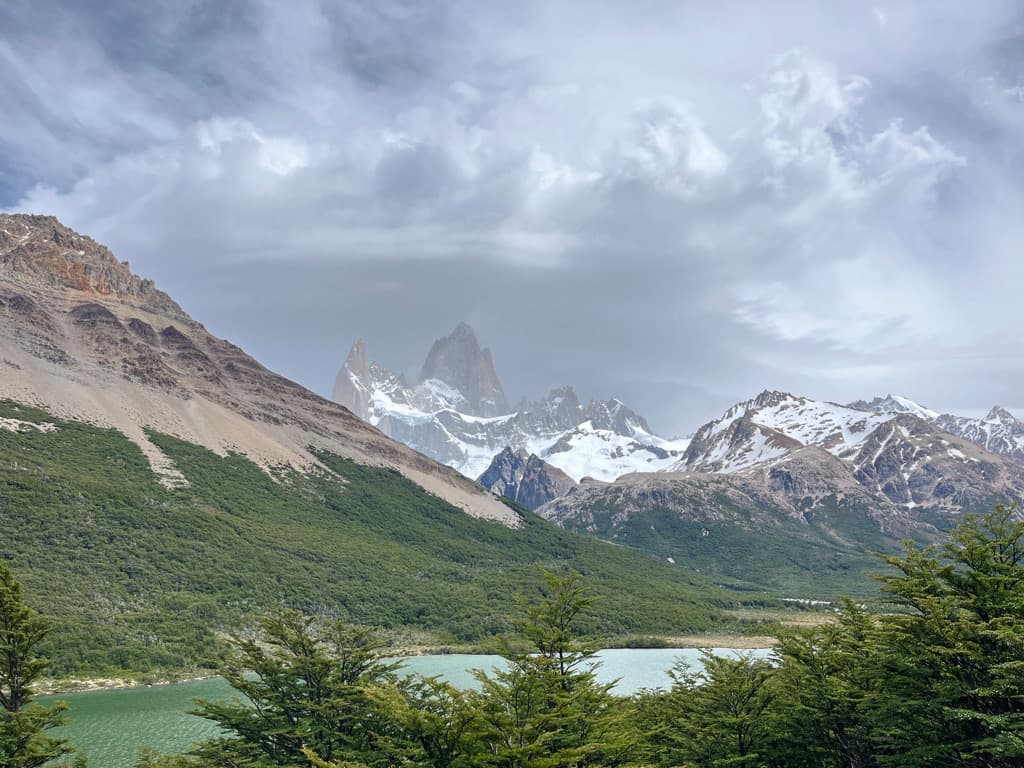 Snow-capped mountain peaks rising above green forest and a turquoise lake beneath a cloudy sky
